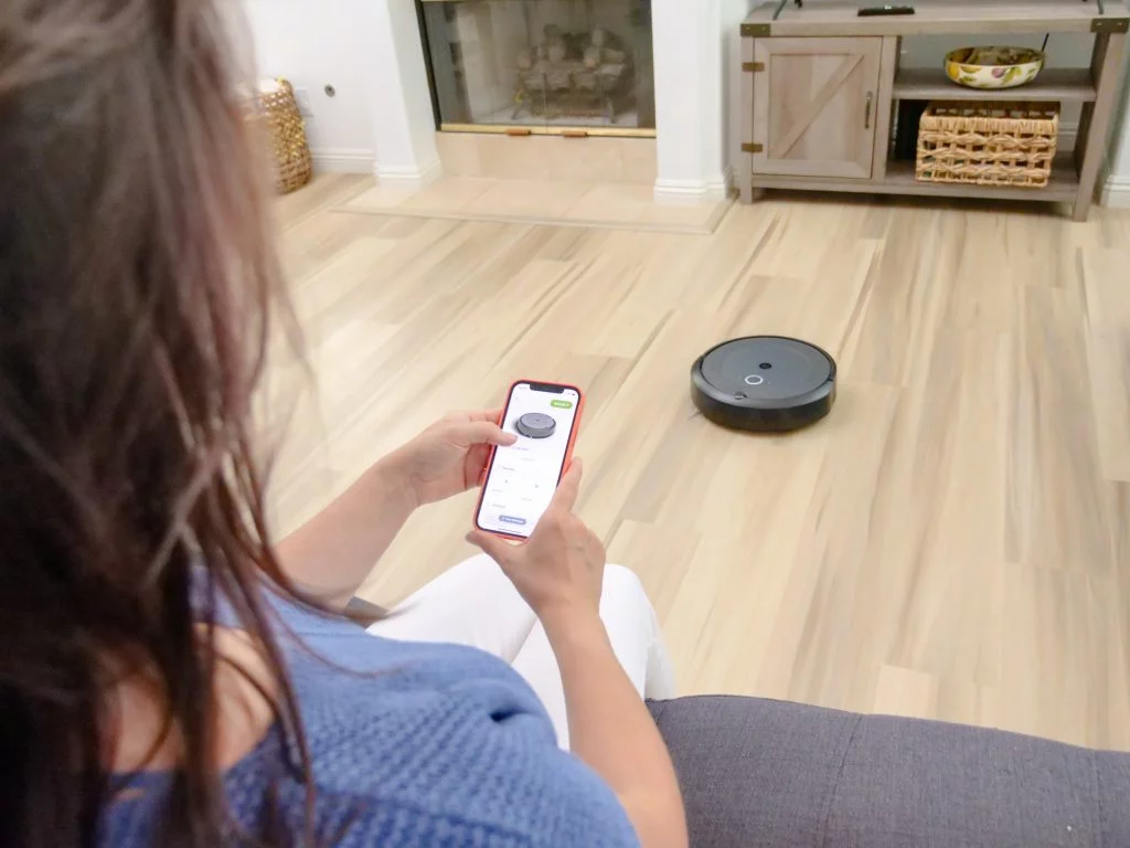 mulher sentada no sofá da sala acompanhando o robô de limpeza, um dos eletrodomésticos inteligentes, pela tela do celular